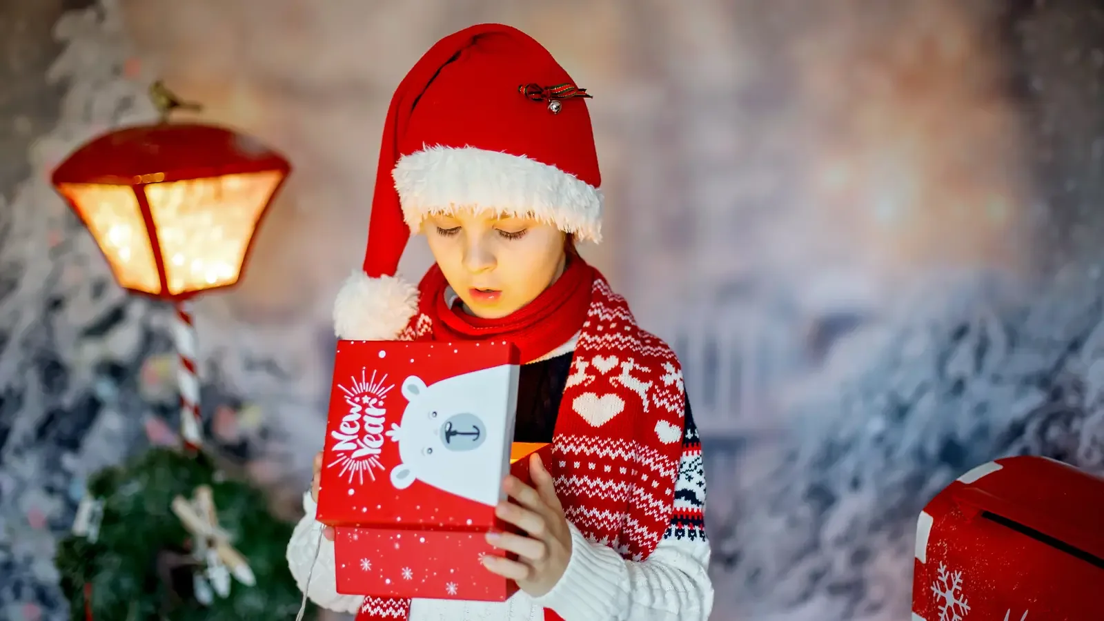 A school-age boy opening a wrapped Christmas present outdoors, surrounded by holiday decoration